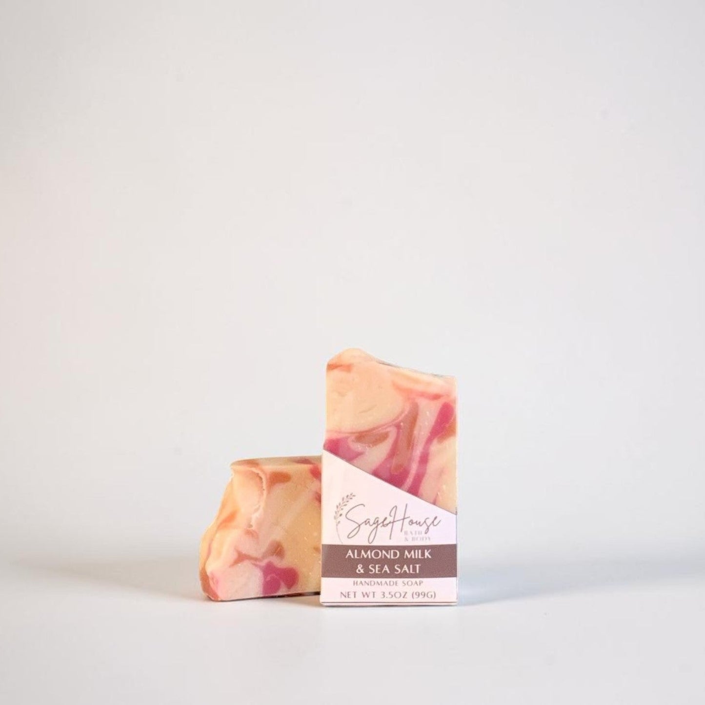A bar of soap with a pink and cream marbled pattern, standing next to its paper packaging sleeve on a light gray background.