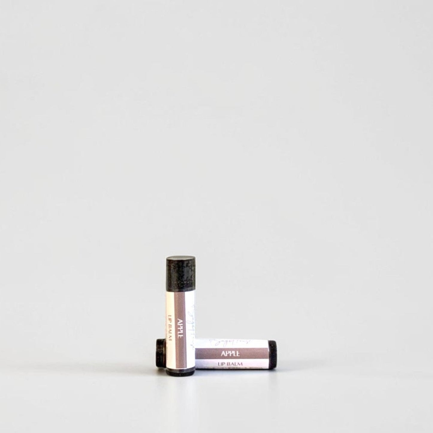 Two slim cylindrical lip balm tubes, one standing upright and one lying horizontally, positioned near the center of a clean white studio background.