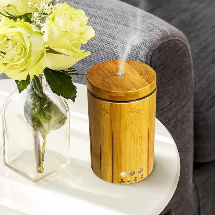 A bamboo diffuser with a wooden top and a glass base, emitting a vapor, placed on a table with a vase of flowers and a couch in the background.