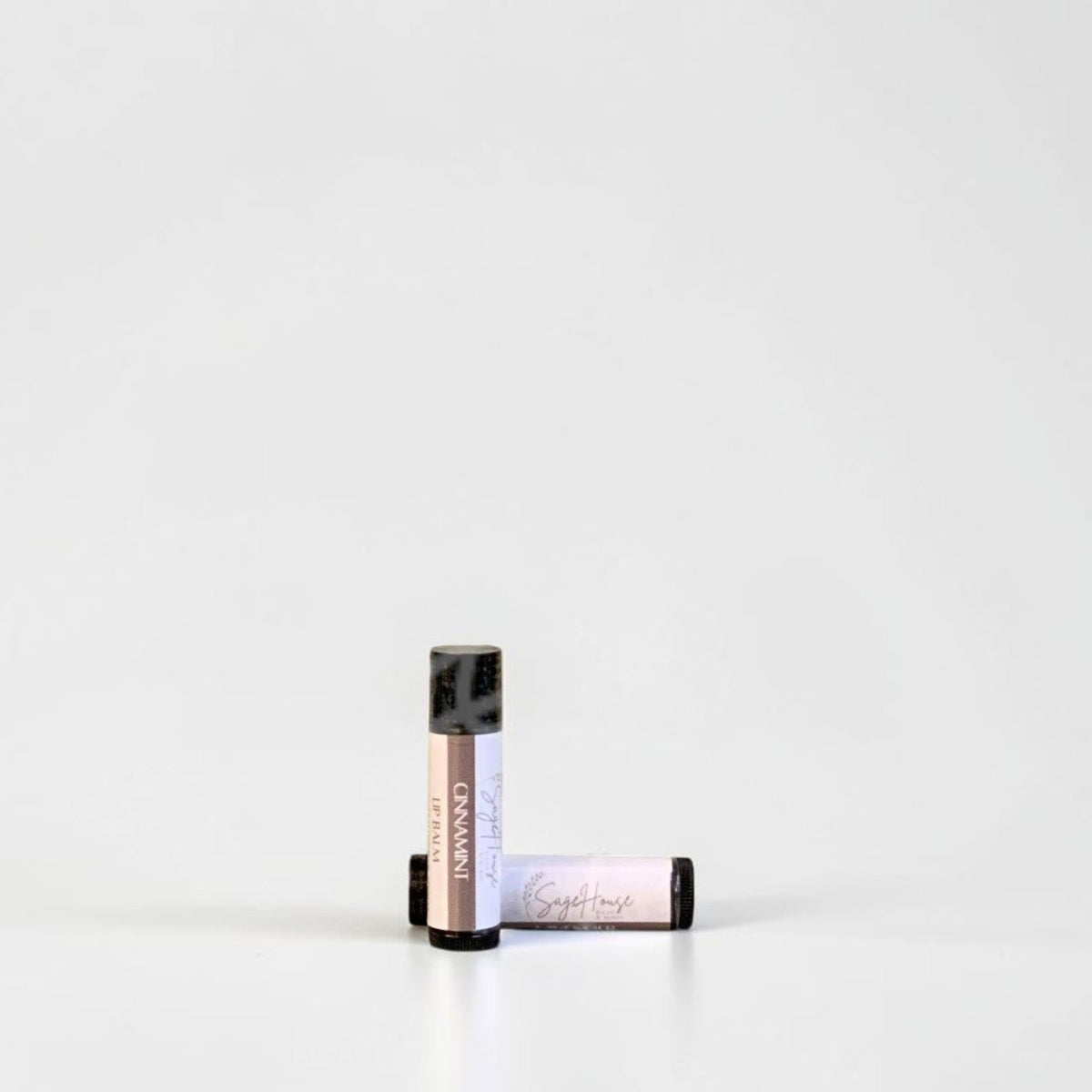 Two slim cylindrical lip balm tubes, one standing upright and one lying horizontally, positioned near the center of a clean white studio background.