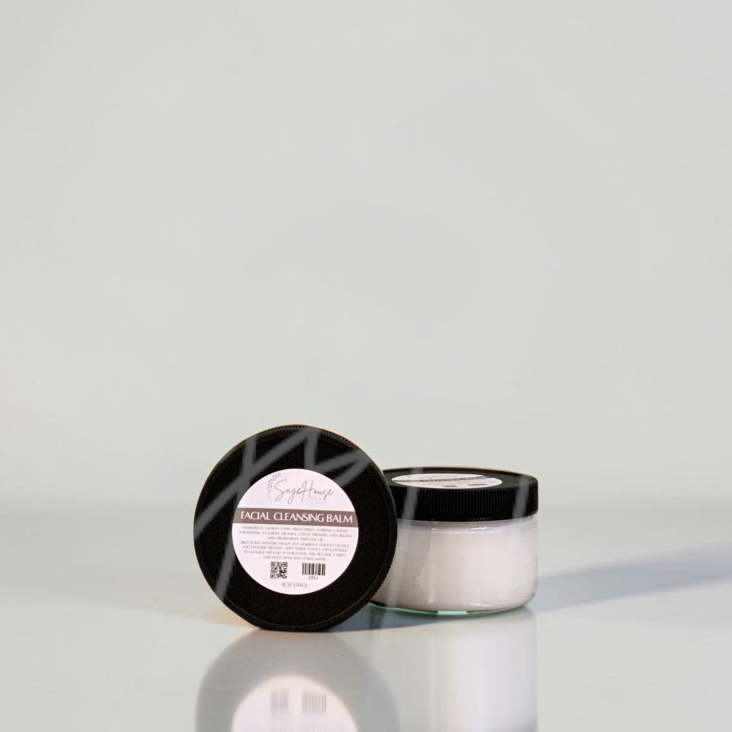 A small cosmetic jar with a black lid and a white label sits on a light gray reflective surface against a matching light gray background.
