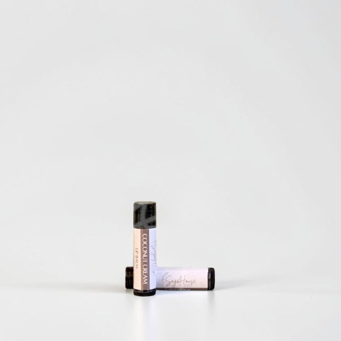 Two slim cylindrical lip balm tubes, one standing upright and one lying horizontally, positioned near the center of a clean white studio background.