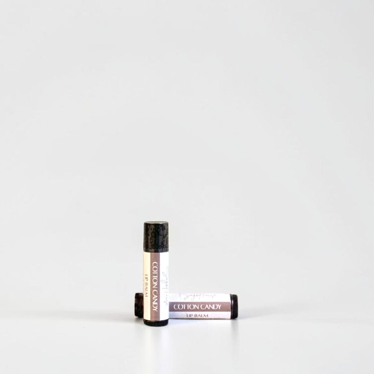 Two slim cylindrical lip balm tubes, one standing upright and one lying horizontally, positioned near the center of a clean white studio background.