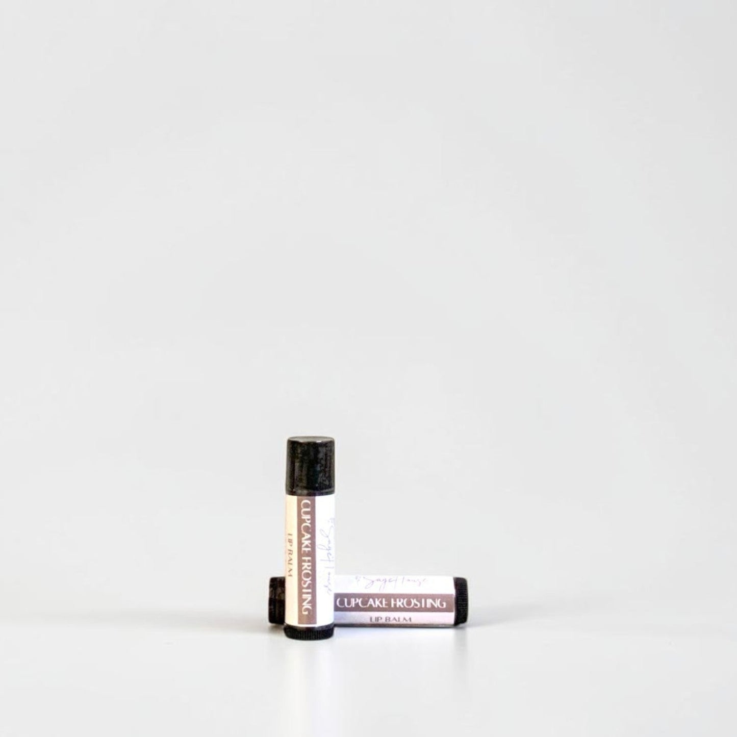Two slim cylindrical lip balm tubes, one standing upright and one lying horizontally, positioned near the center of a clean white studio background.