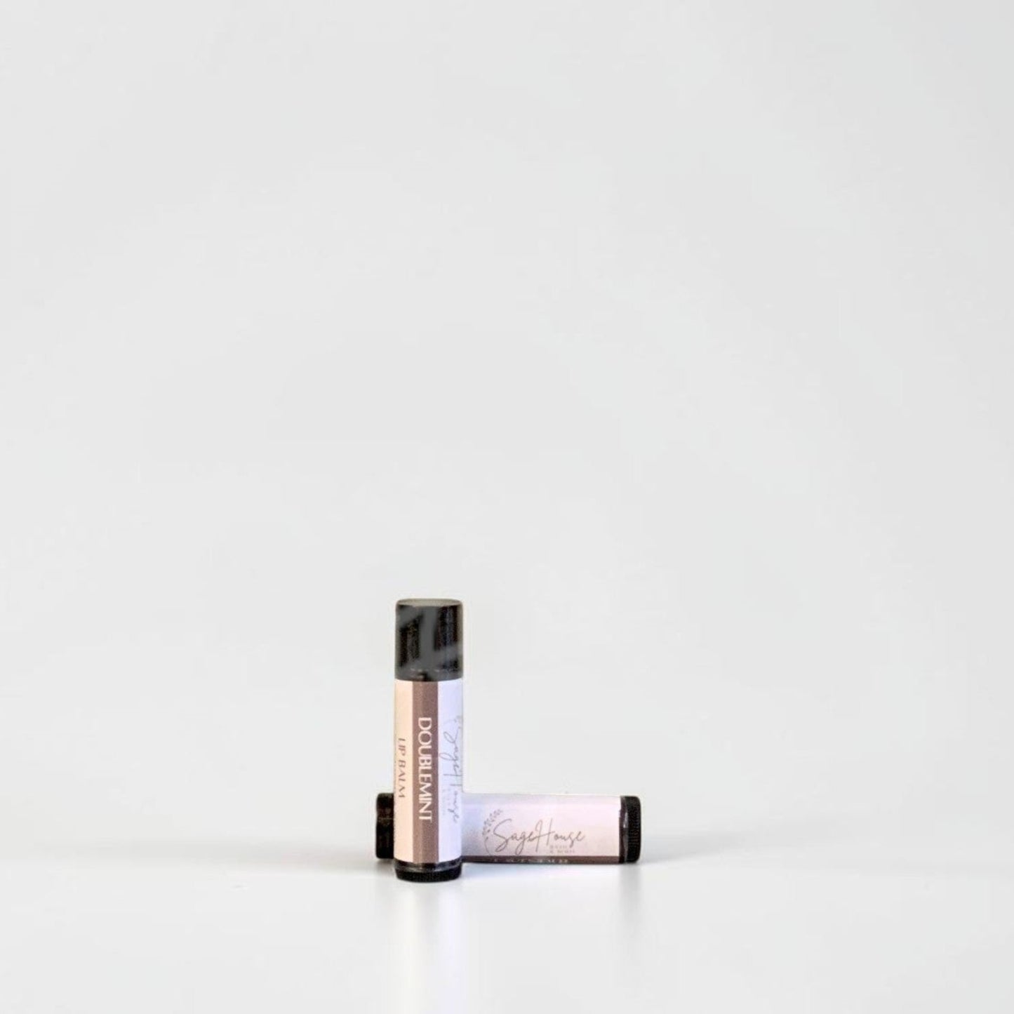 Two slim cylindrical lip balm tubes, one standing upright and one lying horizontally, positioned near the center of a clean white studio background.