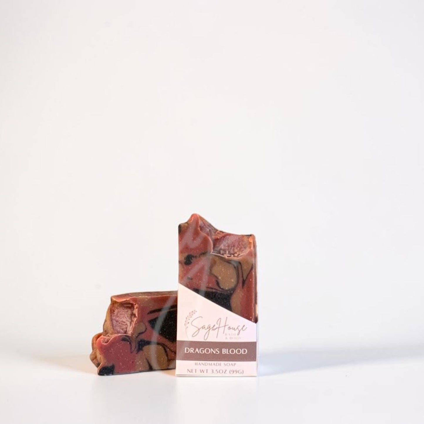 A handmade soap bar with a marbled pattern of reddish-brown, orange, and cream, standing next to its white paper packaging box on a clean white surface.