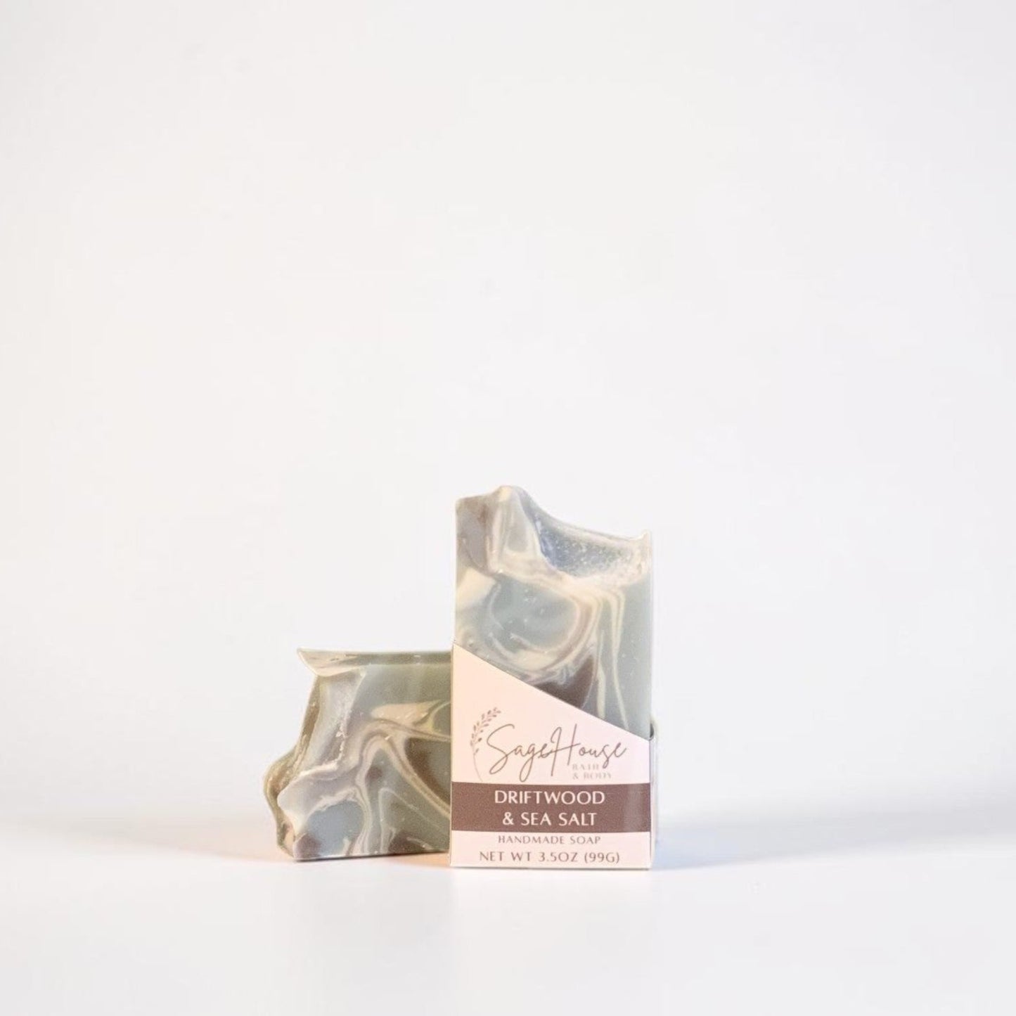 A bar of soap with a light gray and white marbled pattern and a brown label, standing next to its matching paper packaging on a clean white background.
