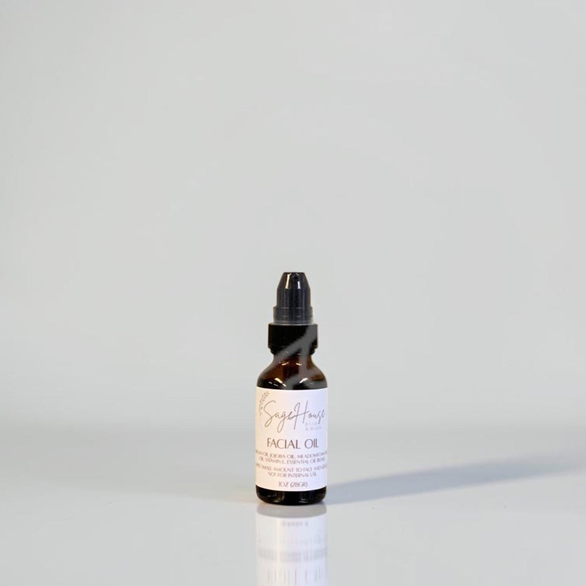 A small amber glass dropper bottle of facial oil with a black dropper cap and a white label, standing on a reflective white surface against a light gray background.