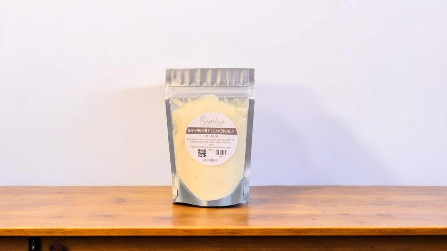 A clear plastic pouch packaging a yellow-colored bath bomb or dust, labeled 'Raspberry Lemonade'. The pouch is placed on a wooden surface.