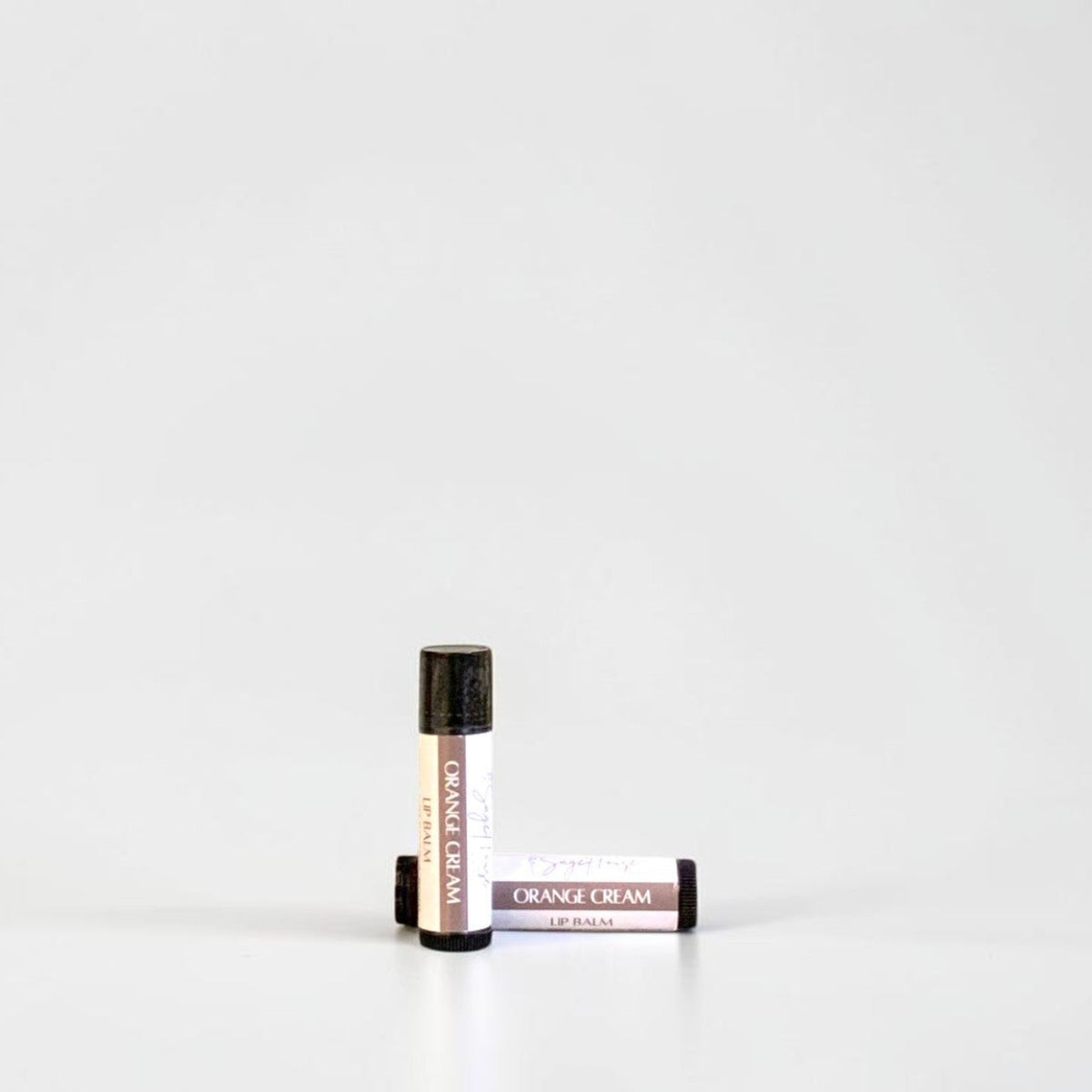 Two slim cylindrical lip balm tubes, one standing upright and one lying horizontally, positioned near the center of a clean white studio background.
