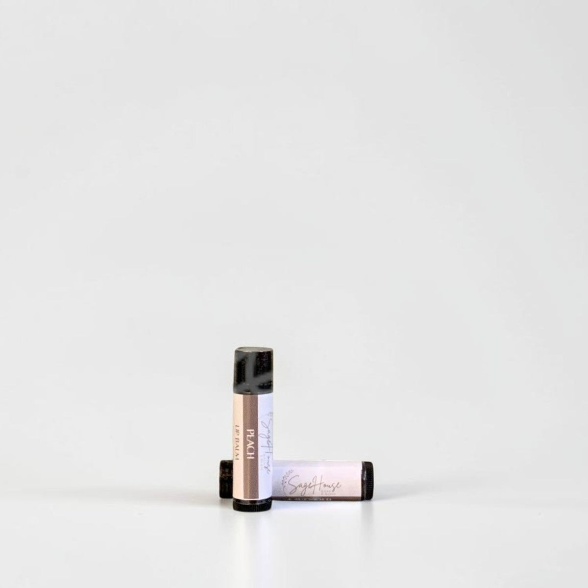 Two slim cylindrical lip balm tubes, one standing upright and one lying horizontally, positioned near the center of a clean white studio background.