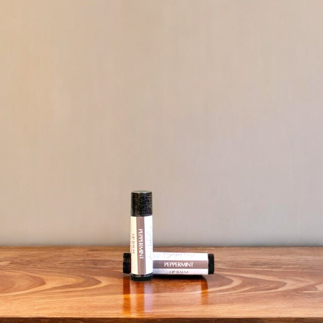 A tube of peppermint lip balm placed upright with a tube lying behind it, on a wooden surface.