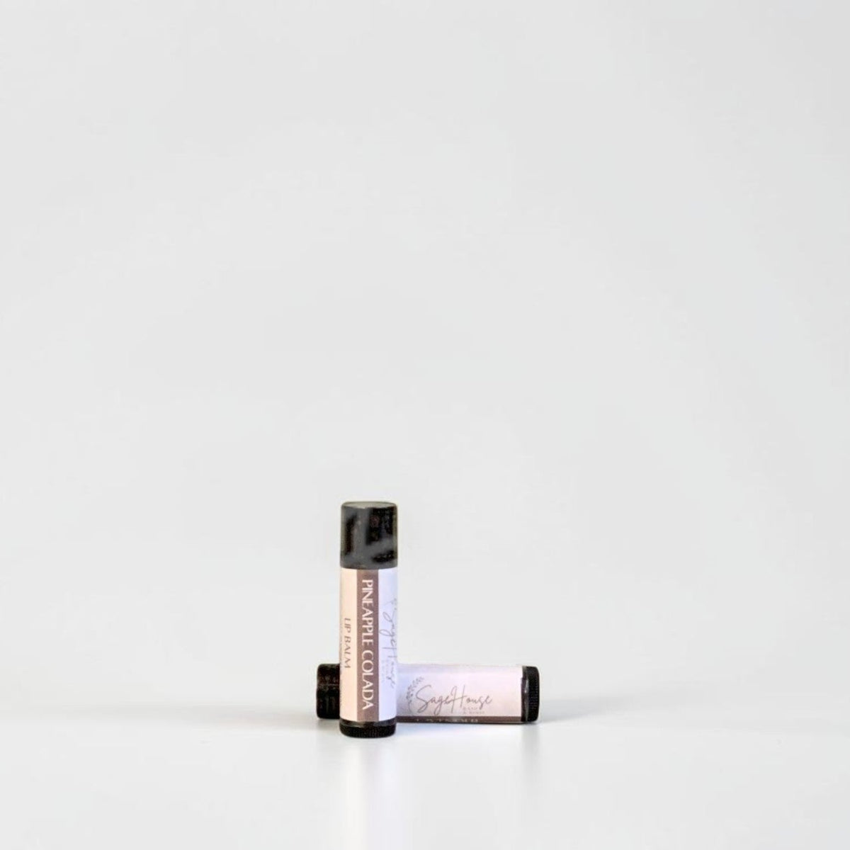 Two slim cylindrical lip balm tubes, one standing upright and one lying horizontally, positioned near the center of a clean white studio background.
