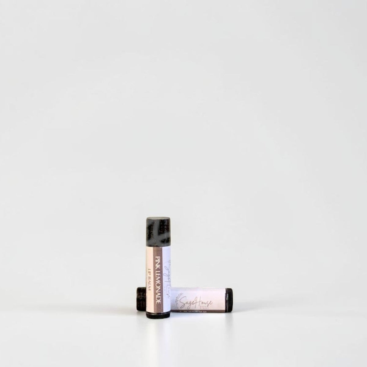 Two slim cylindrical lip balm tubes, one standing upright and one lying horizontally, positioned near the center of a clean white studio background.