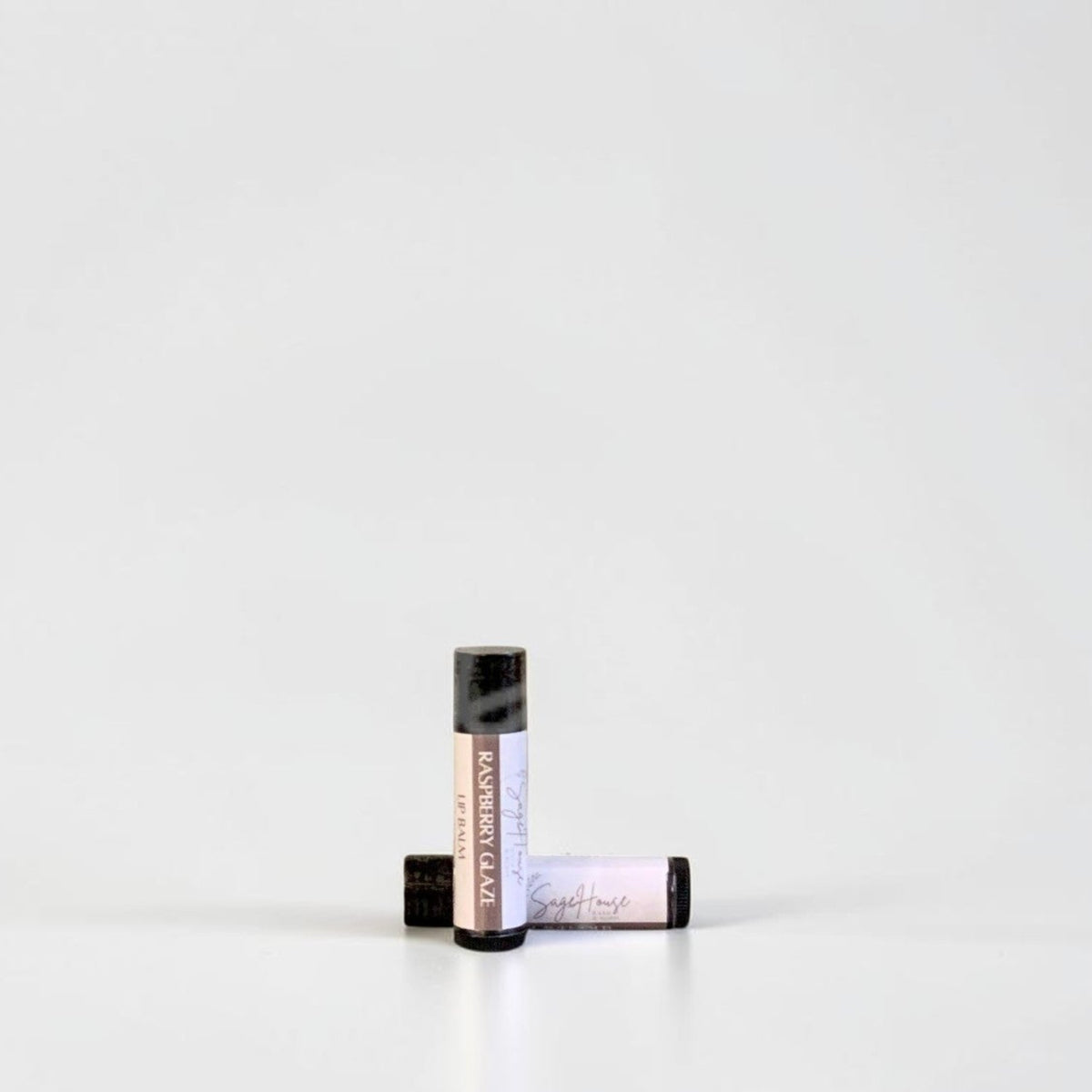 Two slim cylindrical lip balm tubes, one standing upright and one lying horizontally, positioned near the center of a clean white studio background.