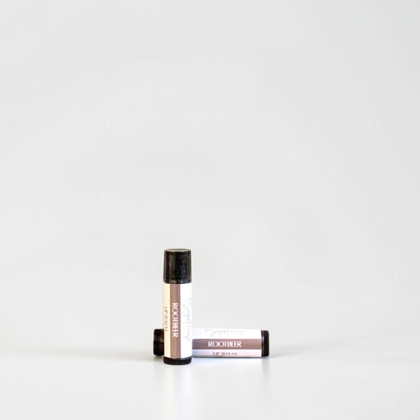 Two slim cylindrical lip balm tubes, one standing upright and one lying horizontally, positioned near the center of a clean white studio background.