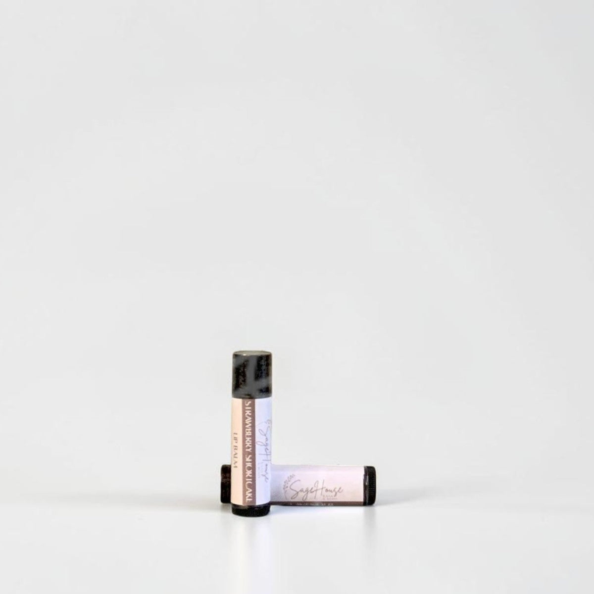 Two slim cylindrical lip balm tubes, one standing upright and one lying horizontally, positioned near the center of a clean white studio background.