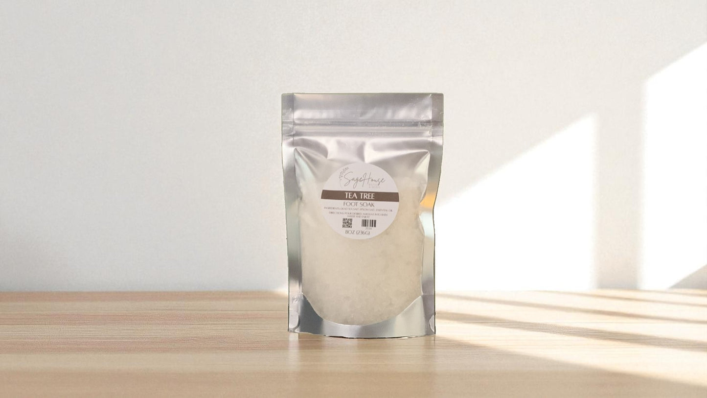 Silver packaging with a clear window of white salt labeled "Tea Tree Foot Soak" on a wood surface.