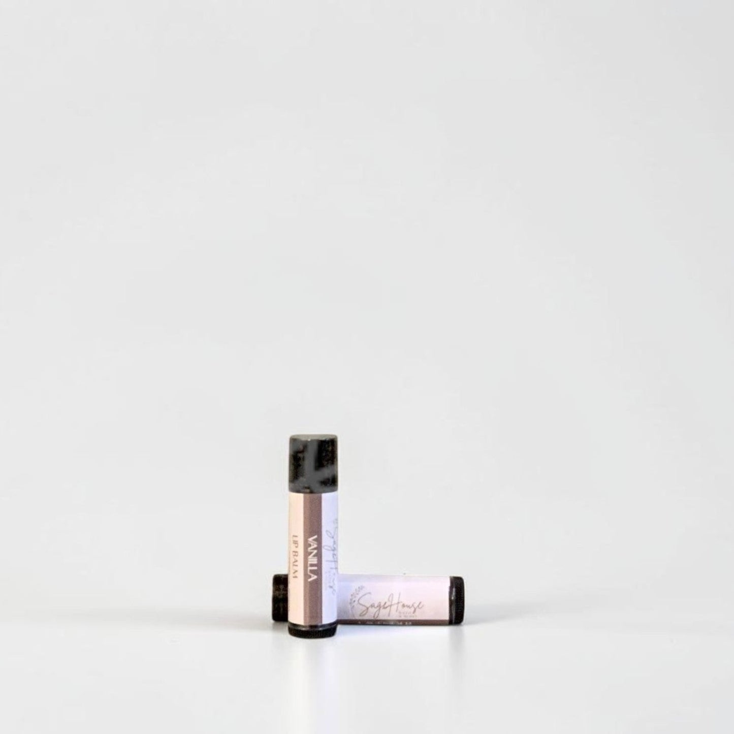 Two slim cylindrical lip balm tubes, one standing upright and one lying horizontally, positioned near the center of a clean white studio background.