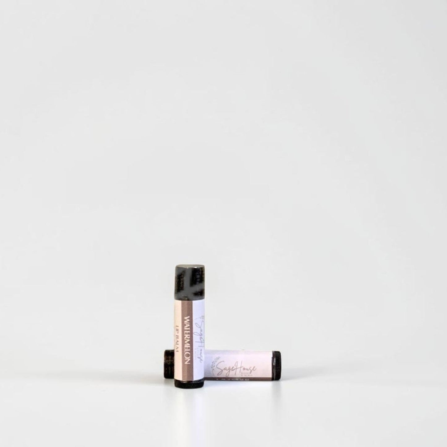 Two slim cylindrical lip balm tubes, one standing upright and one lying horizontally, positioned near the center of a clean white studio background.