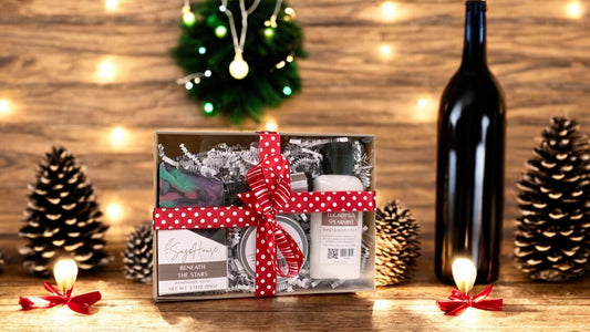 A gift box with a red ribbon, containing a bar of soap, a bottle of lotion, a tube of cuticle oil, and a lip balm, displayed against a Christmas-themed backdrop with lights and pine cones.