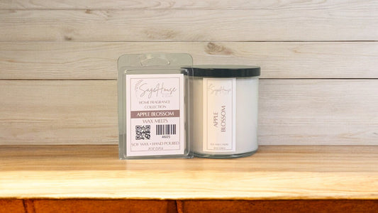 A white soy wax candle in a clear container, next to a clear clamshell wax melt package. placed on a wooden surface, with a label on the front.