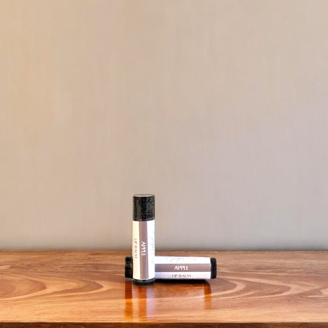 A tube of apple lip balm placed upright with a tube lying behind it, on a wooden surface.