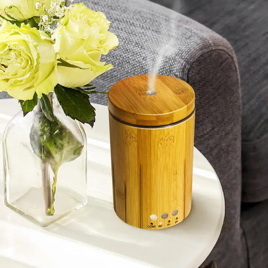 A bamboo diffuser with a wooden top and a glass base, emitting a vapor, placed on a table with a vase of flowers and a couch in the background.