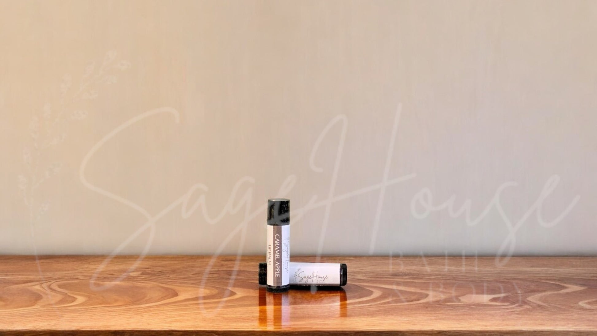 A tube of caramel apple lip balm placed upright with a tube lying behind it, on a wooden surface.