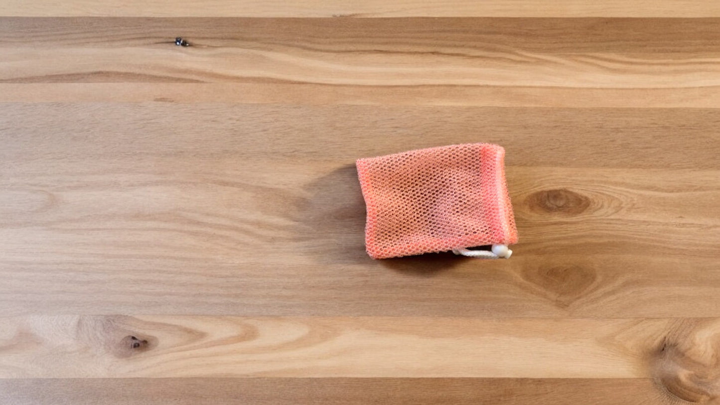 A Soap Scrub Pouch in coral, placed on a wooden surface.