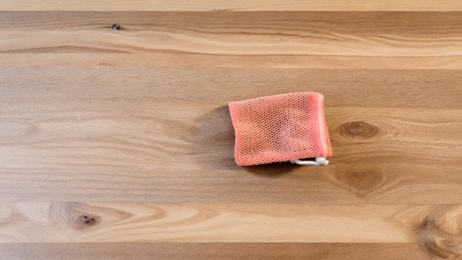 A Soap Scrub Pouch in coral, placed on a wooden surface.