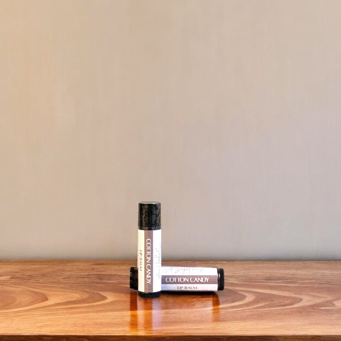 A tube of cotton candy lip balm placed upright with a tube lying behind it, on a wooden surface.