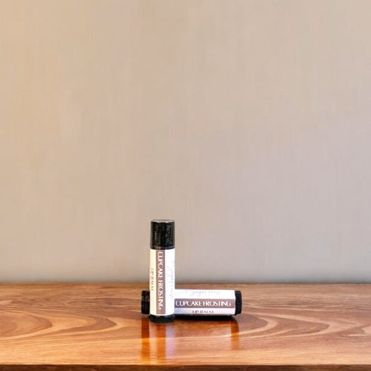 A tube of cupcake frosting lip balm placed upright with a tube lying behind it, on a wooden surface.