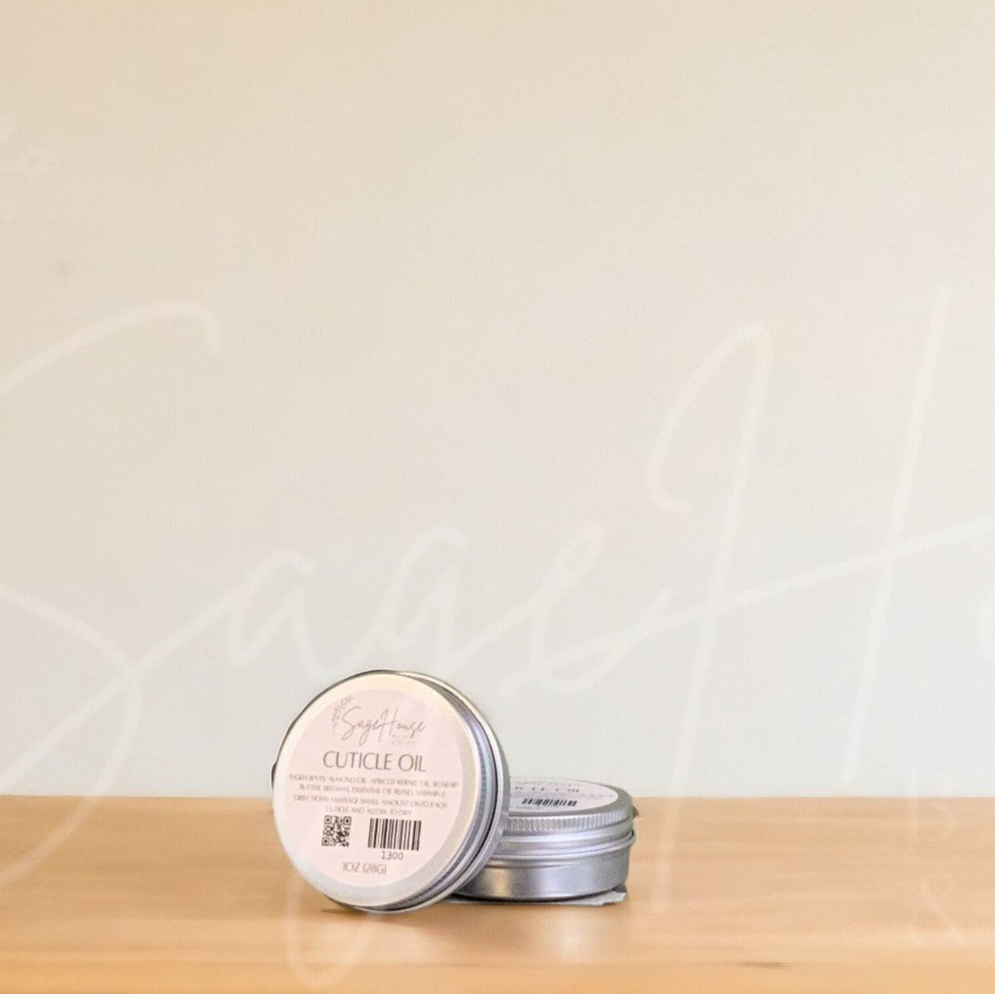 Two small round metal tins of cuticle oil resting on a light wood surface against a warm off-white wall.