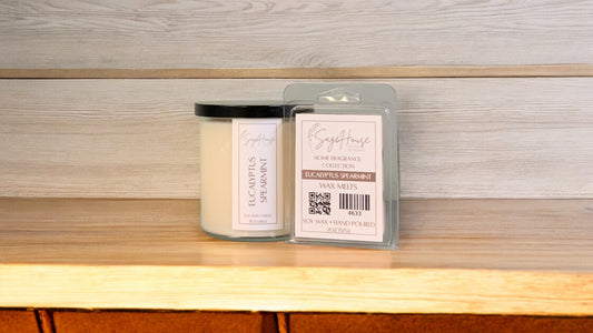 A white soy wax candle in a clear container, next to a clear clamshell wax melt package. placed on a wooden surface, with a label on the front.