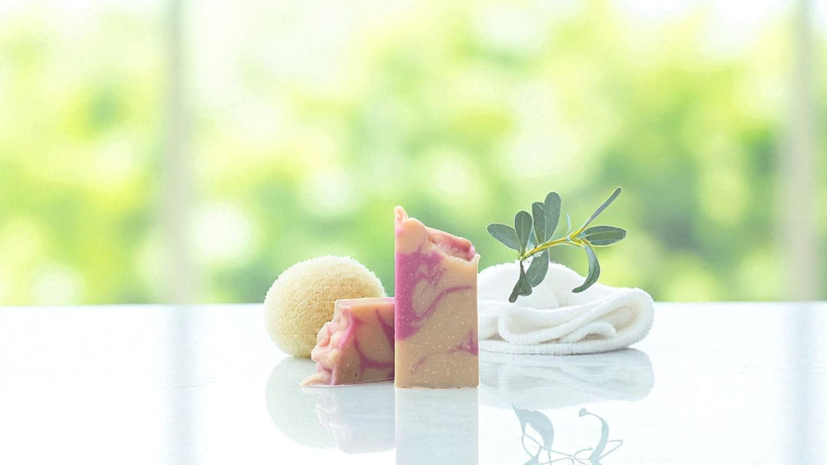 A bar of natural toned soap, with accents of hot pink, surrounded by a sponge on one side, and a towel and sprig of greenery on the other, on a white countertop with a blurred background