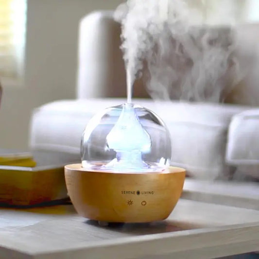 A diffuser with a wooden base and glass globe on top with visible steam coming out the top. On a gray table with a neutral couch in the background