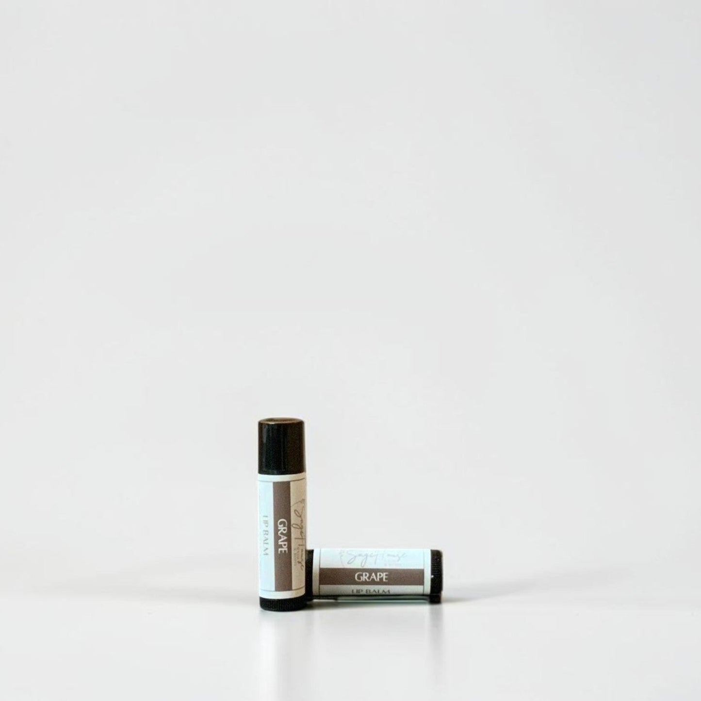 Two slim cylindrical lip balm tubes, one standing upright and one lying horizontally, positioned near the center of a clean white studio background.