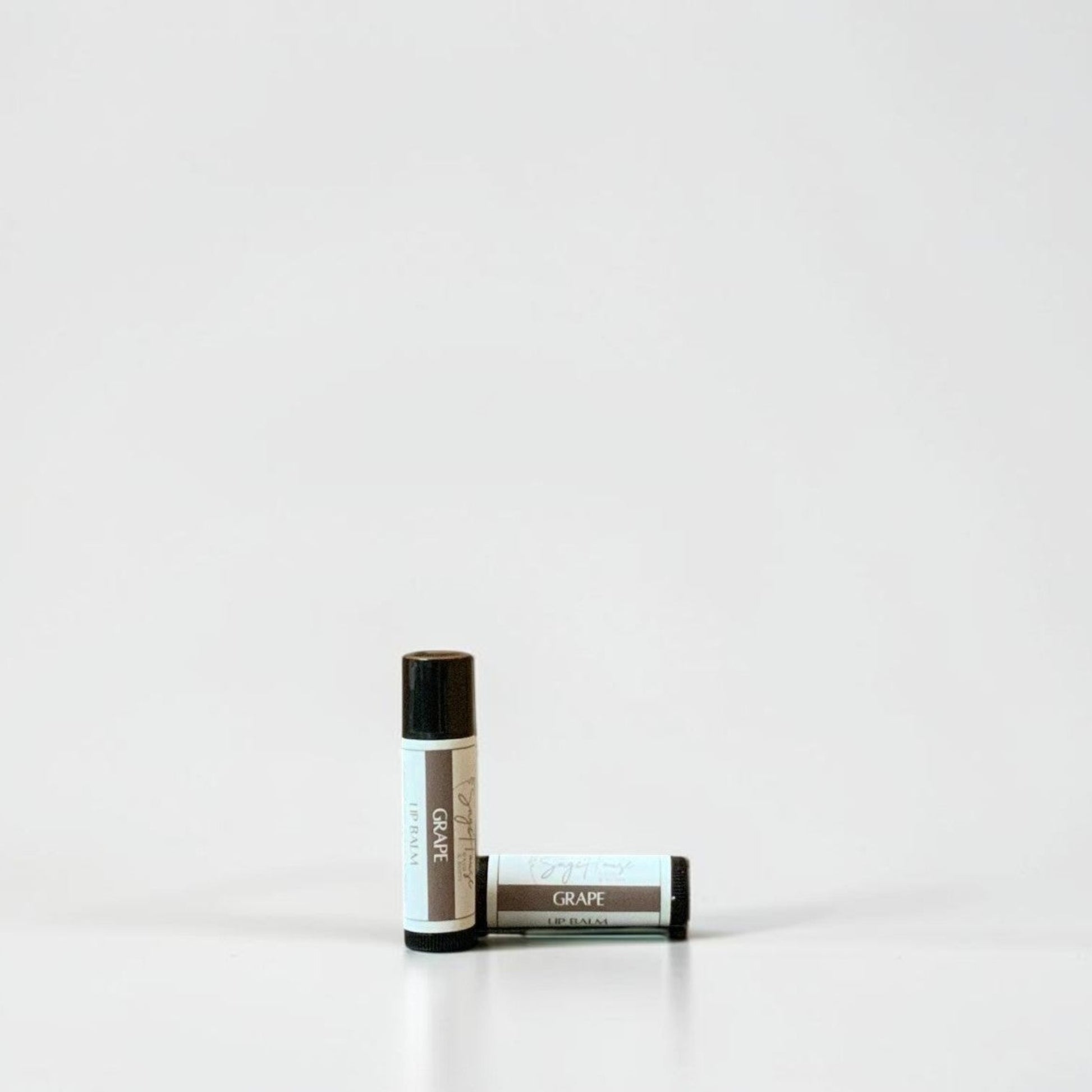 Two slim cylindrical lip balm tubes, one standing upright and one lying horizontally, positioned near the center of a clean white studio background.
