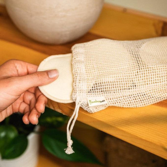 A hand holding a beige hemp facial round, with another round visible inside a beige cotton mesh wash bag.
