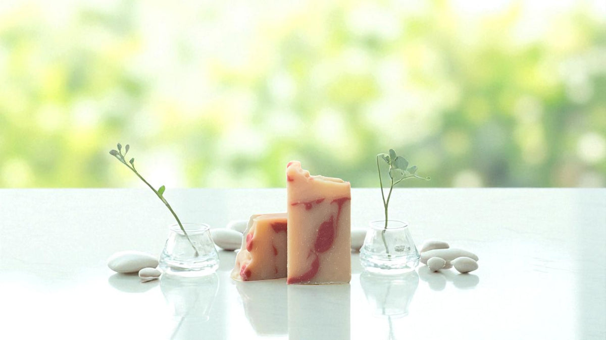 A bar of peach toned soap, with accents of red, surrounded by stones and clear vases containing small sprigs of greenery, on a white countertop with a blurred background