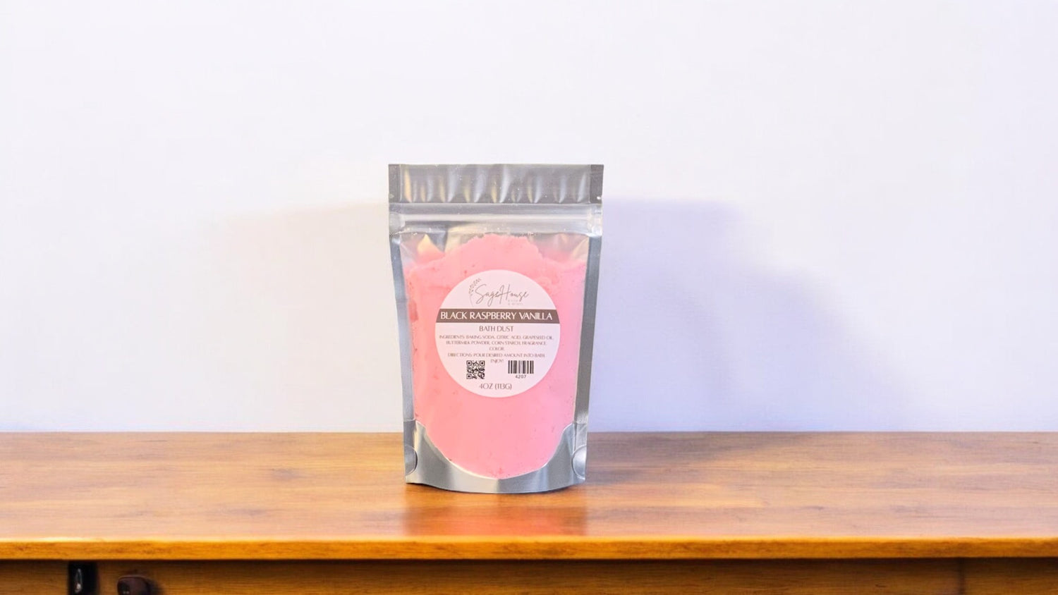 A clear plastic pouch packaging a pink-colored bath bomb or dust, labeled 'black raspberry vanilla'. The pouch is placed on a wooden surface.