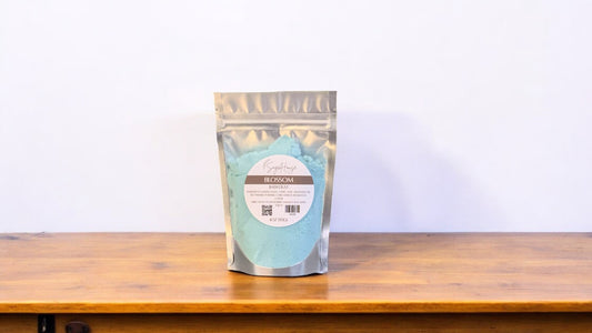 A clear plastic pouch packaging a teal-colored bath bomb or dust, labeled 'blossom'. The pouch is placed on a wooden surface.