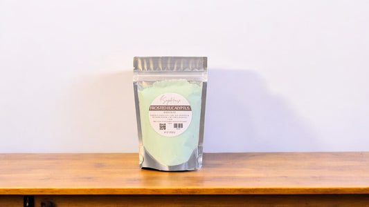 A clear plastic pouch packaging a light green-colored bath bomb or dust, labeled 'frosted eucalyptus'. The pouch is placed on a wooden surface.
