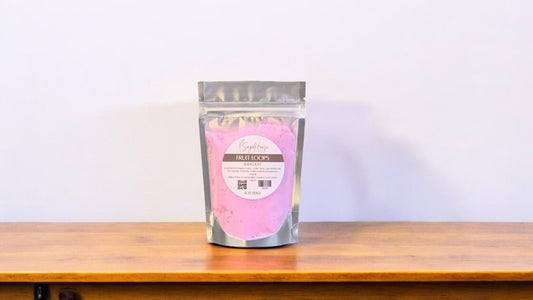 A clear plastic pouch packaging a purple-colored bath bomb or dust, labeled 'fruit loops'. The pouch is placed on a wooden surface.