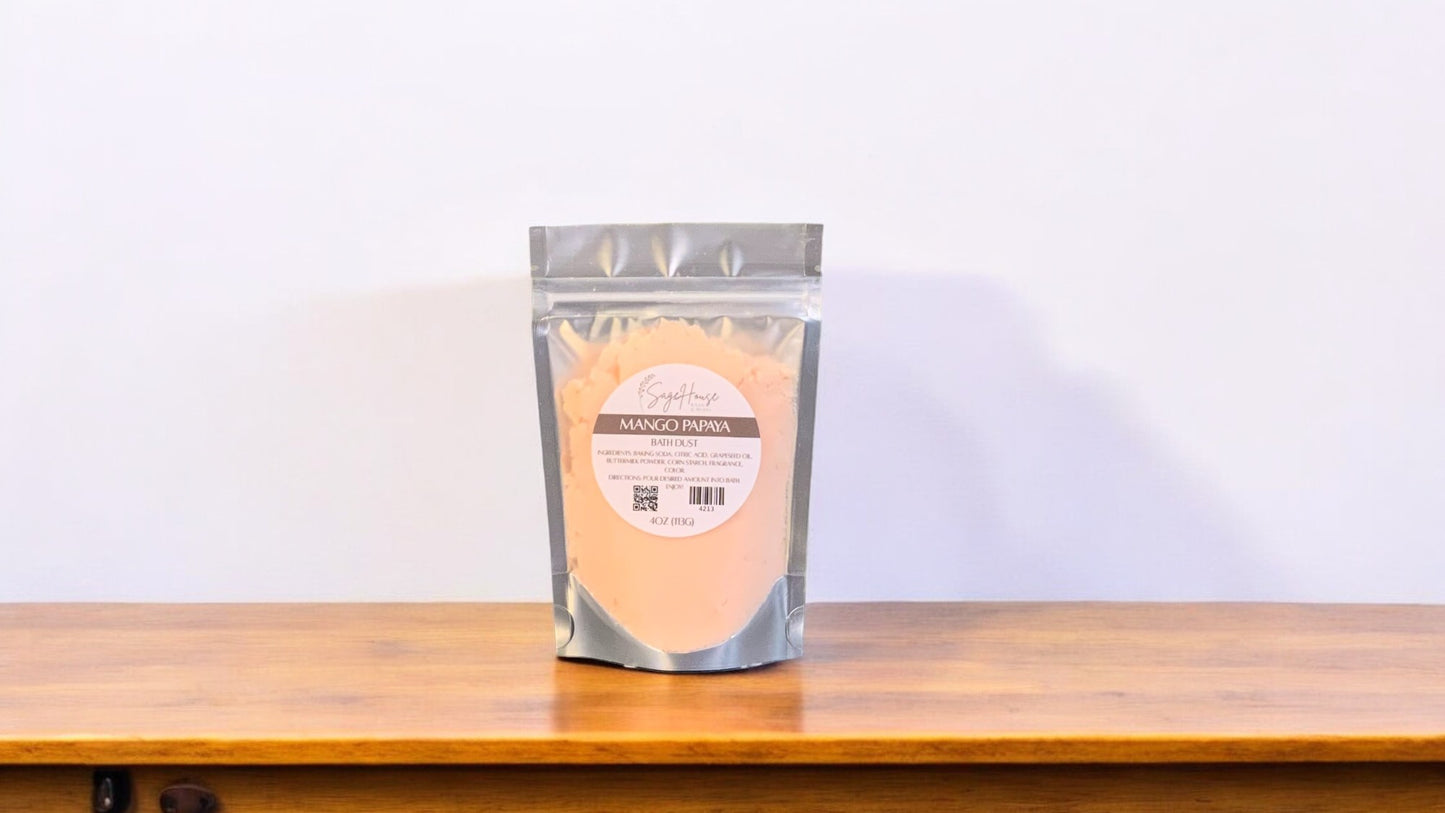 A clear plastic pouch packaging a peach-colored bath bomb or dust, labeled 'mango papaya'. The pouch is placed on a wooden surface.