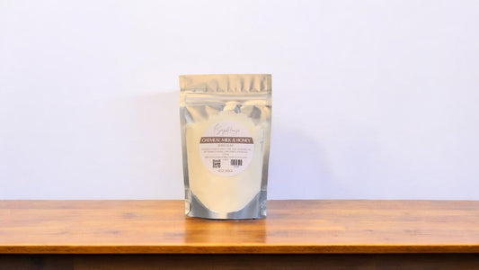 A clear plastic pouch packaging a beige-colored bath bomb or dust, labeled 'oatmeal milk and honey'. The pouch is placed on a wooden surface.