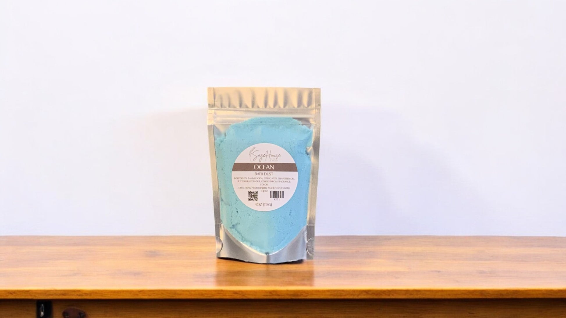 A clear plastic pouch packaging a blue-colored bath bomb or dust, labeled 'ocean'. The pouch is placed on a wooden surface.