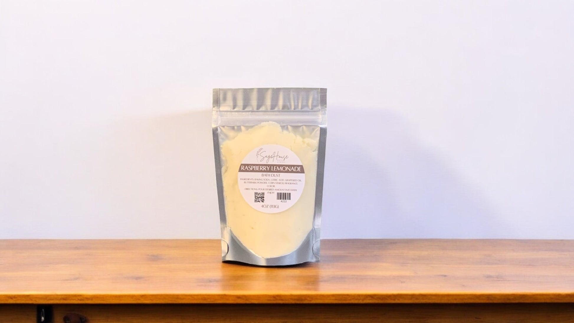 A clear plastic pouch packaging a yellow-colored bath bomb or dust, labeled 'Raspberry Lemonade'. The pouch is placed on a wooden surface.