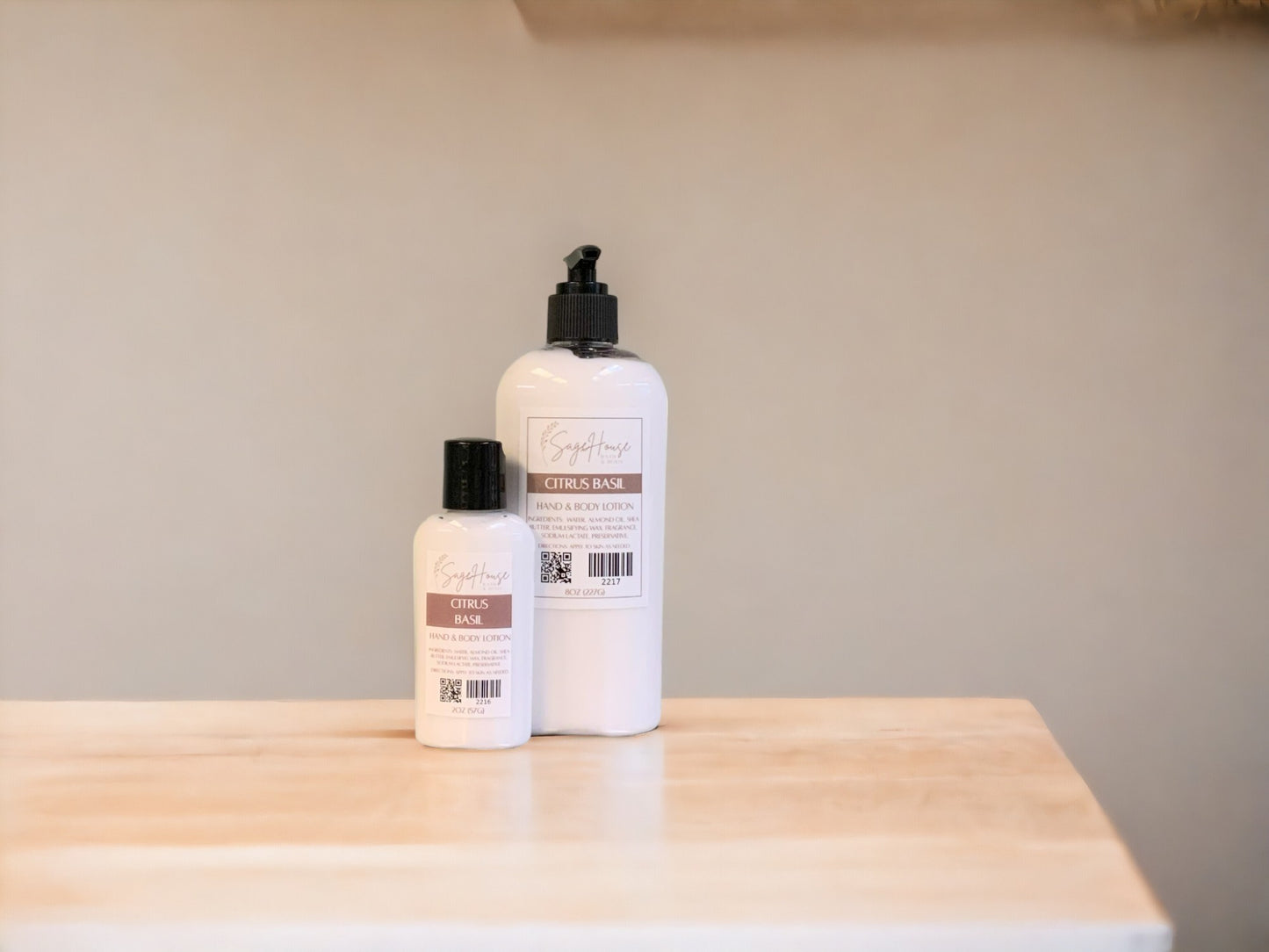 A small bottle of lotion, next to a large bottle of lotion, sitting on a light wood surface, with a tan background. 
